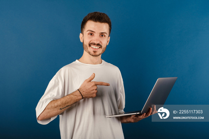 Young man smiling confident wearing white tee posing isolated over blue background smiling and pointing with index finger to the laptop pc computer.