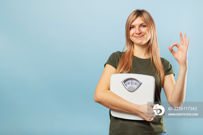 Young blonde woman holding white scale on blue background and doing OK sign