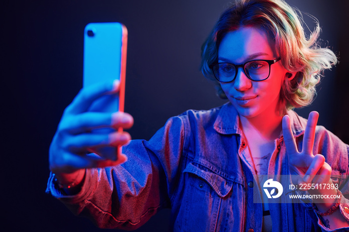 Portrait of young alternative girl in glasses with green hair that making selfie by phone in red and blue neon light in studio