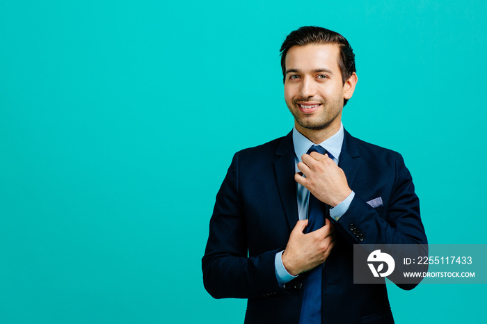 Portrait of a young entrepreneur business man fixing his tie,  isolated on blue studio background