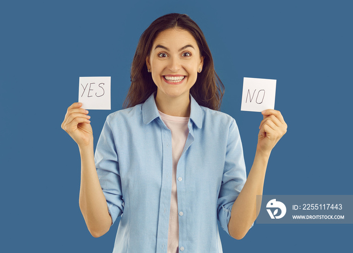 Doubted happy brunette woman with two cards yes and no looking at camera on blue background. Laughing puzzled excited woman with face full of doubt looking at camera. Trying to make a good choice.