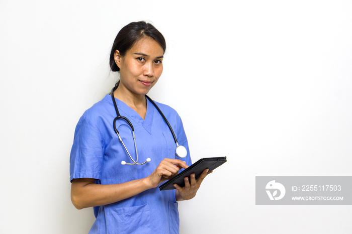 Woman physical therapist in blue uniform and stethoscope typing on tablet computer. Stand in front of white wall.