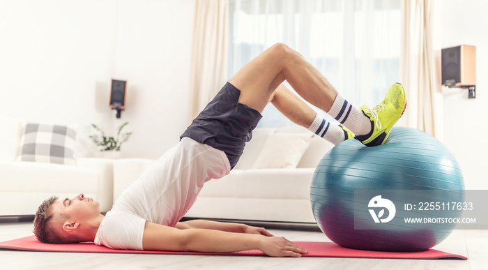 Young male working out with a blue fit ball at home on a red mat, exercising glutes, legs and core of the body