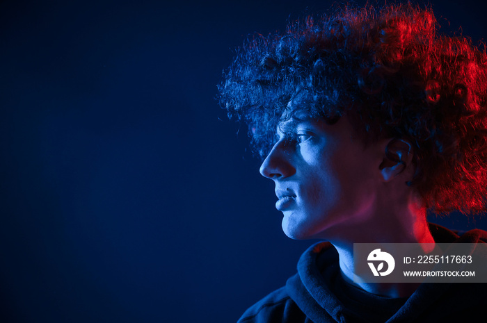 View from the side. Young man with curly hair is indoors illuminated by neon lighting