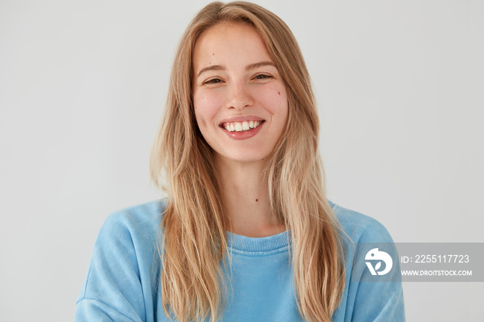 Portrait of happy lovely female has broad smile, being in good mood after party with friends, has long hair, wears casual oversized sweater, isolated over white background. Positive emotions concept