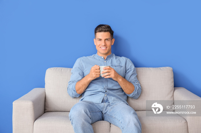 Young man with tea relaxing on sofa near color wall