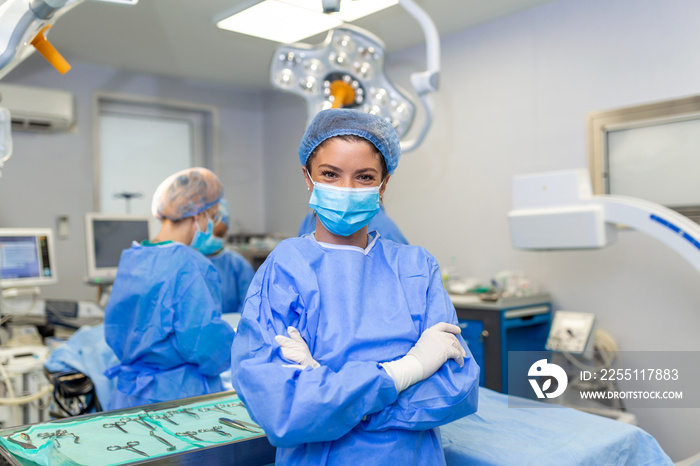 Portrait of smiling surgeon in hospital. Female healthcare worker is wearing scrubs. She is standing with arms crossed against lights.