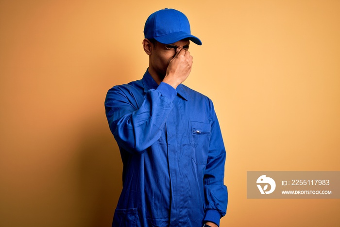 Young african american mechanic man wearing blue uniform and cap over yellow background tired rubbing nose and eyes feeling fatigue and headache. Stress and frustration concept.