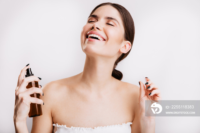 Satisfied brunette girl after shower sprays mist for body on skin. Woman smiling on isolated background