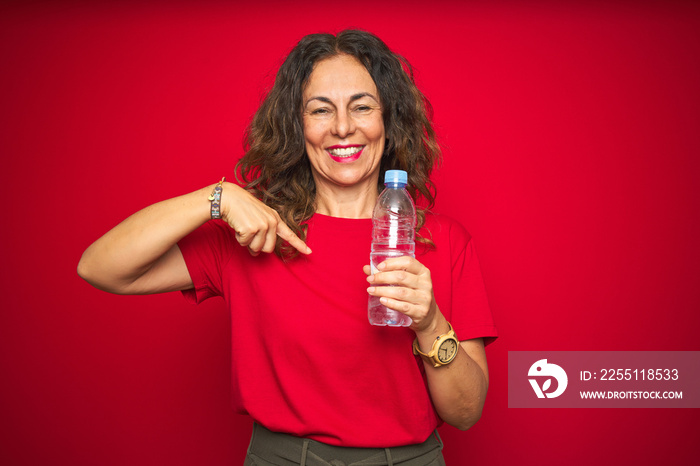 Middle age senior woman holding plastic water bottle over red isolated background with surprise face pointing finger to himself