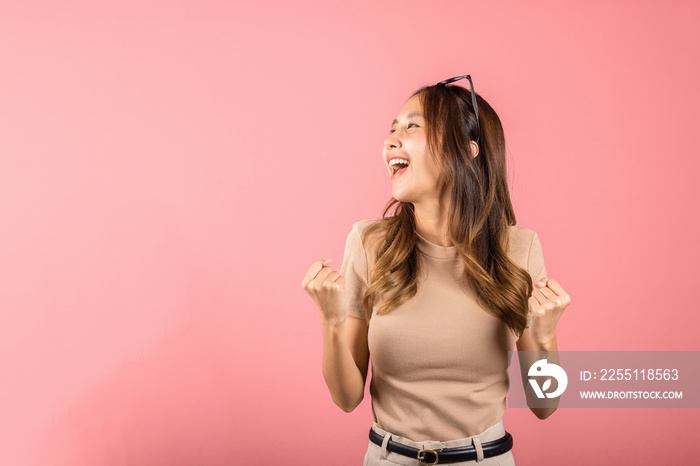 Happy Asian portrait beautiful cute young woman wear glasses makes raised hand up celebrating her winning success gesture, studio shot isolated pink background, Female excited say yes! with copy space