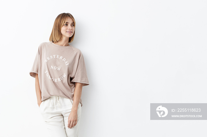 Beautiful young female with boobed hair, wearing loose shirt and white trousers, modeling against white background, looking aside while waiting for her boyfriend, going to have walk in with him