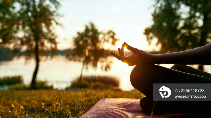 Cropped shot of a woman doing yoga outdoors near lake or river at sunrise in the morning, sitting in lotus pose on the background of nature