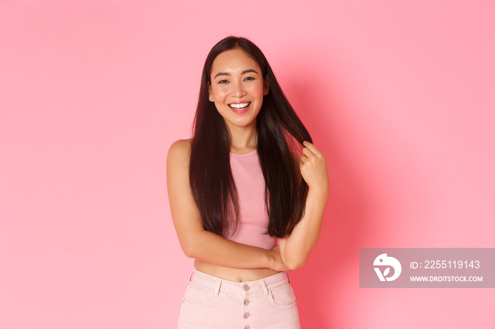 Lifestyle, beauty and women concept. Portrait of stylish carefree asian girl playing with hair while talking, smiling pleased, having lively conversation with girlfriends over pink background