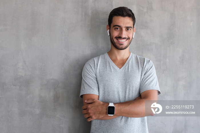 Portrait of smiling handsome man in t shirt and smart watches, standing with crossed arms against gray textured wall with copy space