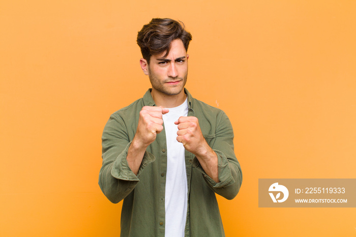 young handsome man looking confident, angry, strong and aggressive, with fists ready to fight in boxing position against orange background