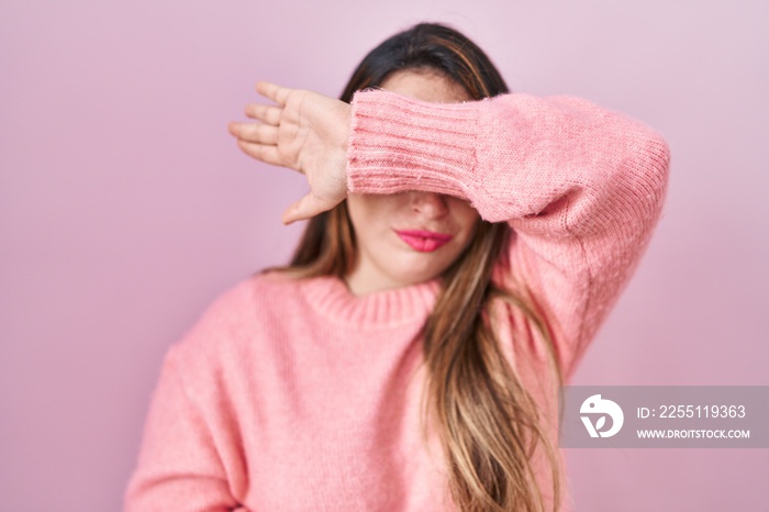 Young hispanic woman standing over pink background covering eyes with arm, looking serious and sad. sightless, hiding and rejection concept