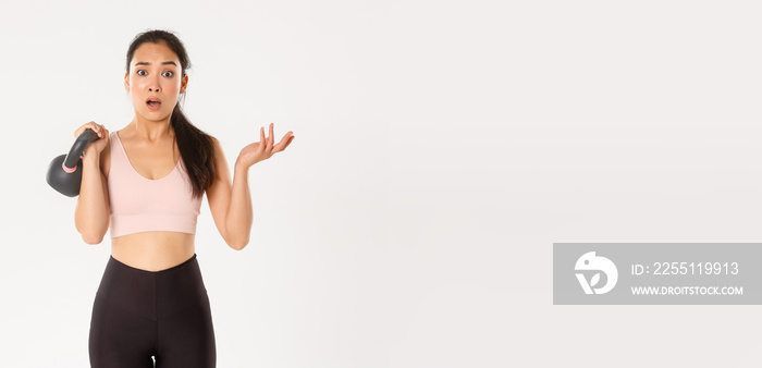 Sport, wellbeing and active lifestyle concept. Confused asian fitness girl, female athlete lifting kettlebell and look puzzled, consulting coach during training session, standing white background