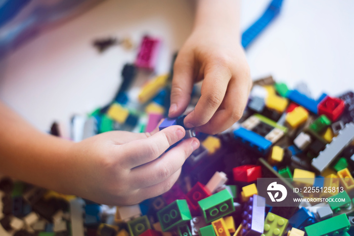 Hands of a child constructing Lego against the background of a heap of multicolored constructor blocks.