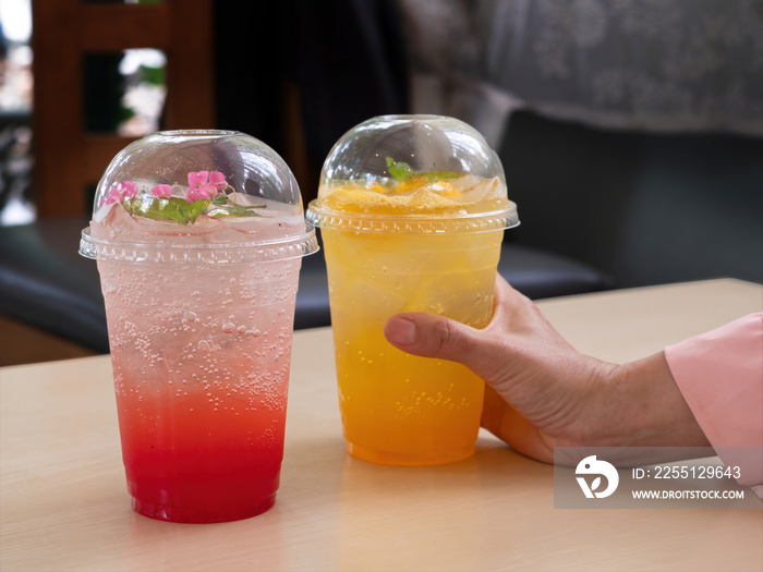 Woman’s hand holding one take away glass filled with mango soda drinks. Two glasses of colorful refreshing drinks, Very Berry Soda and Mango Passion Soda on the table in coffee shop.