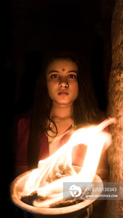 beautiful Indian girl in old stepwell wearing traditional Indian red saree, gold jewellery and bangles holding fire plate. Maa Durga shoot concept. Indian girl dancer in the posture of Indian dance