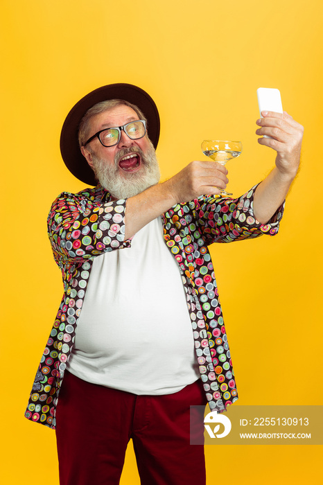 Time to drink and selfie. Portrait of senior hipster man in eyewear isolated on yellow studio background. Tech and joyful elderly lifestyle concept. Trendy colors, forever youth. Copyspace for your ad