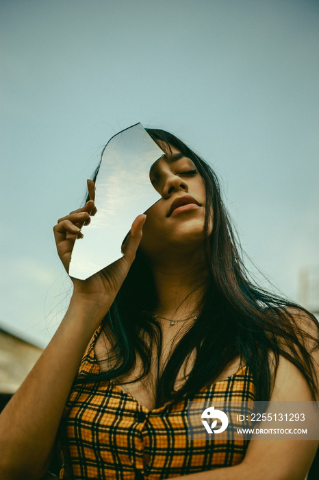 Girl in orange print dress holding piece of broken mirror reflecting sky clouds in front of her face