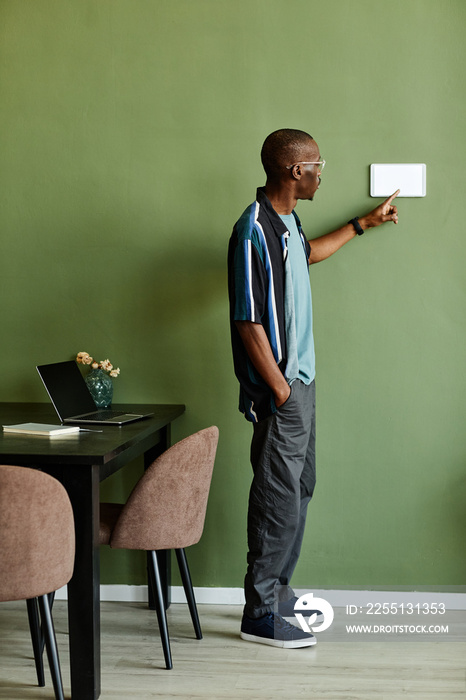 Vertical full length portrait of African American man using smart home control panel in minimal modern interior