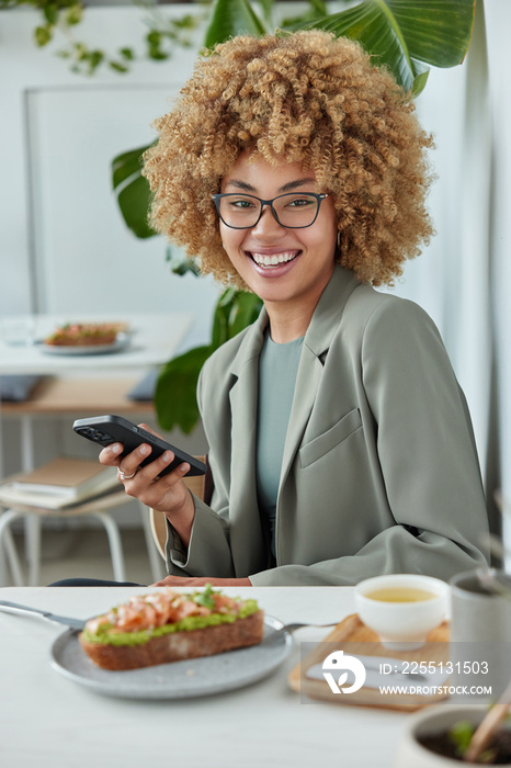 Young positive woman uses cellphone for online networking connected to wireless internet wears spectacles and formal clothes has lunch in cozy cafeteria rests after work. People technology lifestyle