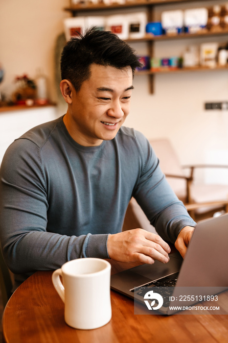 Happy adult asian man smiling and using laptop in cafe indoors