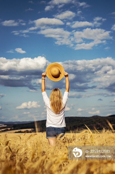 Happy woman holding straw hat is standing in wheat field. Enjoyment of summer