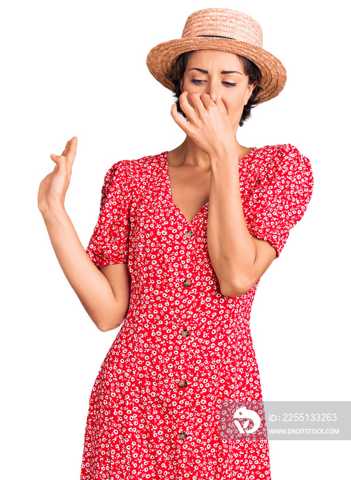 Young beautiful woman wearing summer hat smelling something stinky and disgusting, intolerable smell, holding breath with fingers on nose. bad smell