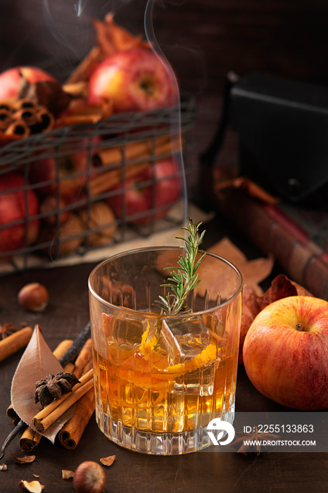 Glass of alcoholic whiskey drink with a branch of rosemary with smoke and apples, cinnamon sticks. Concept of magical autumn relaxed and cozy atmosphere.