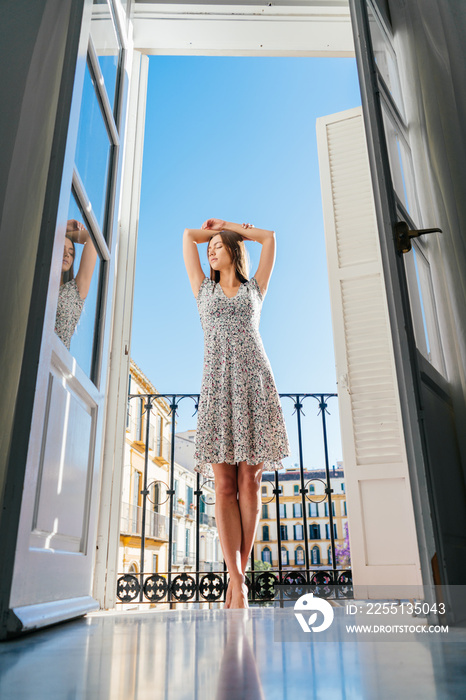 a barefoot girl in a light dress rose on tiptoe and stretches her arms behind her head while standing on the hotel balcony on the background of European buildings and blue sky
