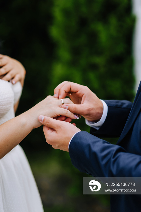 The groom in a blue suit puts a gold ring on the finger of the bride’s hand at the ceremony. Wedding photography close-up of the newlyweds, portrait, macro.