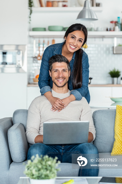 Happy beautiful couple using their laptop to searching voyage while looking at camera sitting on the couch at home.