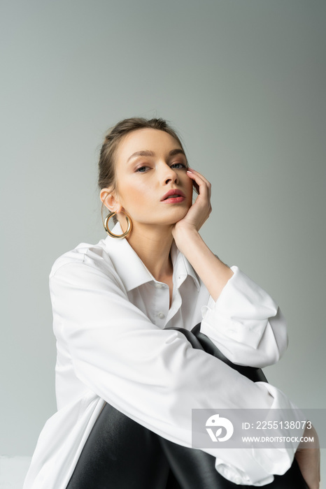 stylish woman in white oversize shirt holding hand near face and looking at camera on grey background.