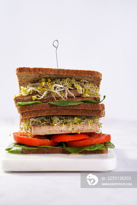 Tasty vegan sandwiches with whole brown protein bread, tofu cheese, tomato, spinach and microgreens sprouts stacked on top of each other, white background