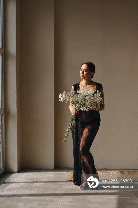 Studio portrait of brunette pregnant woman in black peignoir holding a bouquet of gypsophila in her hands and smiling