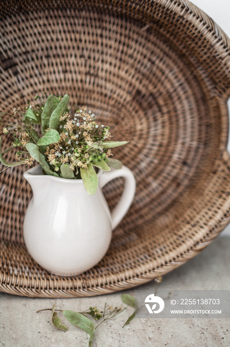 Ceramic beige jug with dry lime blossoms against the background of an oval rattan tray closeup.