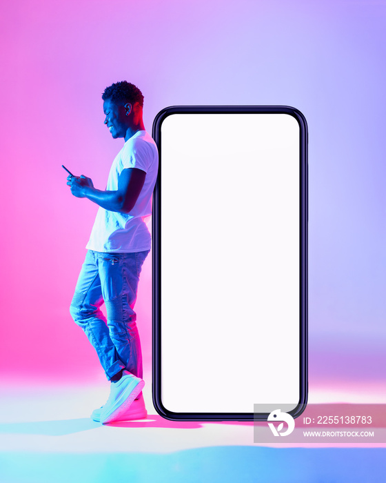 Full length of young black man using mobile phone while leaning on huge smartphone with empty white screen in neon light
