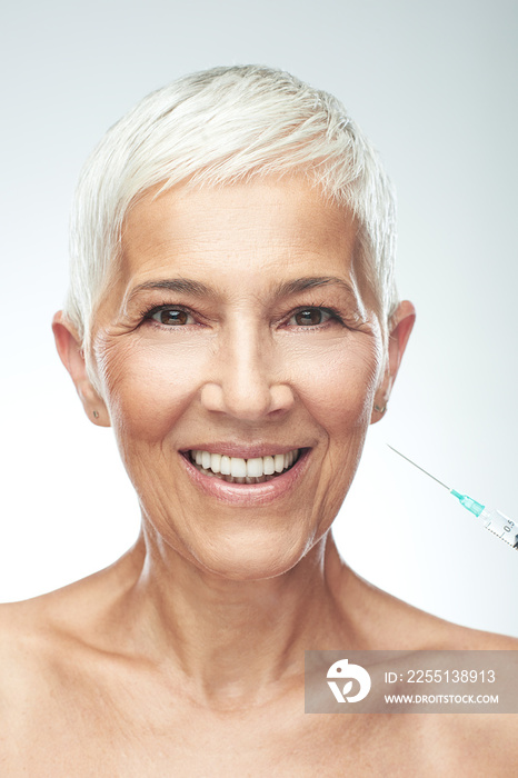 Beautiful smiling Caucasian senior woman standing in front of gray background and taking a shot of anti age product. Beauty photography.