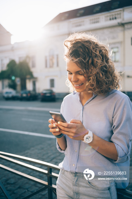 Happy curly young woman texting and smiling on the smart phone