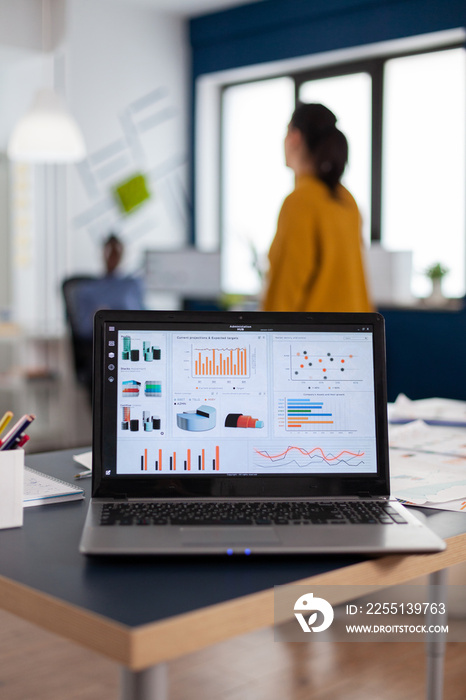 Close up of laptop with graphs, charts of marketing agency. Workplace area in business center with multi ethnic employees in it, shot of room with modern furniture and blue wall.