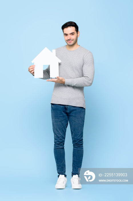 Handsome smiling Caucasian man holding house model cutout standing in light blue isolated studio background