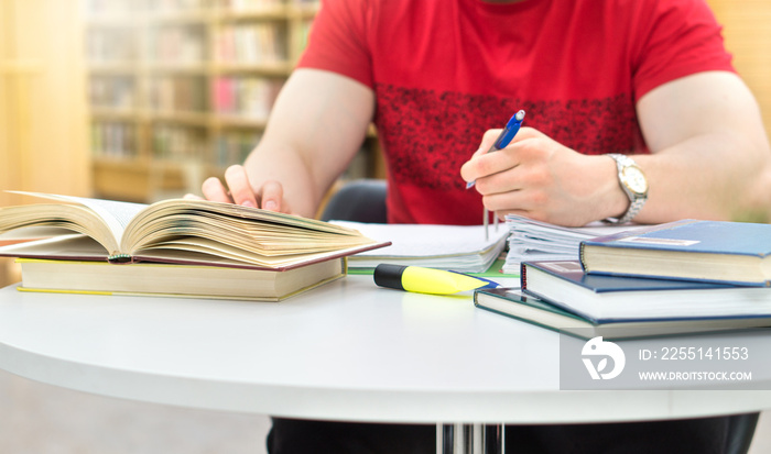 Young athletic man and student studying and writing notes in public or school library in college or university. Stack and pile of books, pen and paper on table.