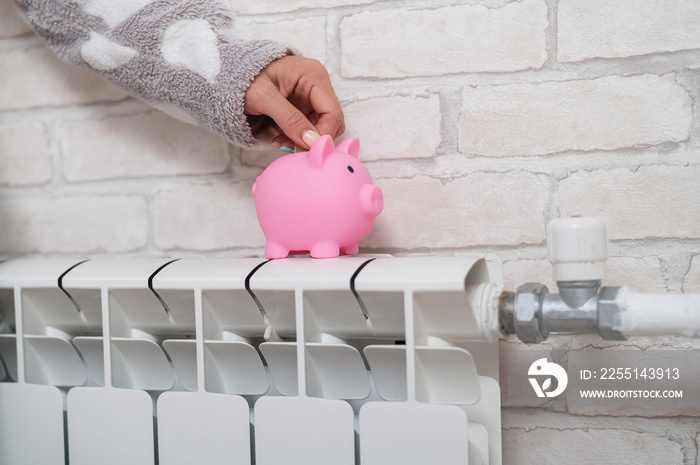 Female hand putting coin into piggy bank which standing on heating radiator with temperature regulator on brick wall background