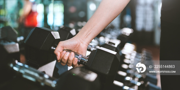 Closeup of hands of young fit and healthy female in sportswear lifting single hand weights from rack with multiple dumbbells while practicing workout for arms in modern gym and fitness club