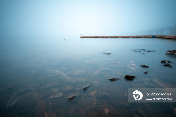 Wooden jetty on a foggy river with stones in the water on a foreground. Minimalist long exposure landscape.