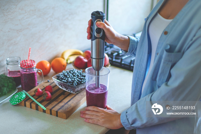 Woman cooking fresh blueberry smoothie with hand blender at kitchen at home. Healthy eating and berry drink